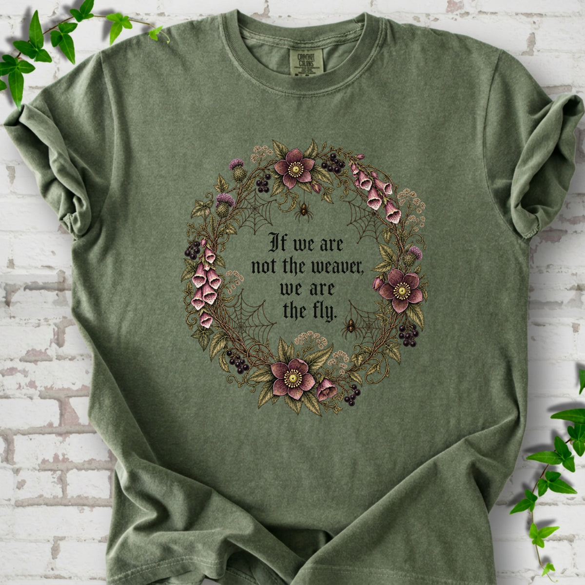 Hemp T-shirt flat-lay mockup with a gothic floral wreath, spider webs and "If we are not the weaver, we are the fly" lettering.