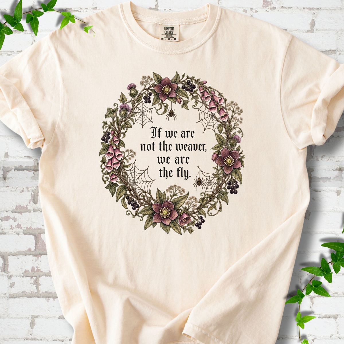 Ivory T-shirt flat-lay mockup with a gothic floral wreath, spider webs and "If we are not the weaver, we are the fly" lettering.
