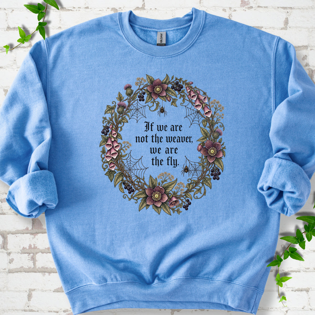 Carolina Blue sweatshirt flat-lay mockup with a gothic floral wreath, spider webs and "If we are not the weaver, we are the fly" lettering.
