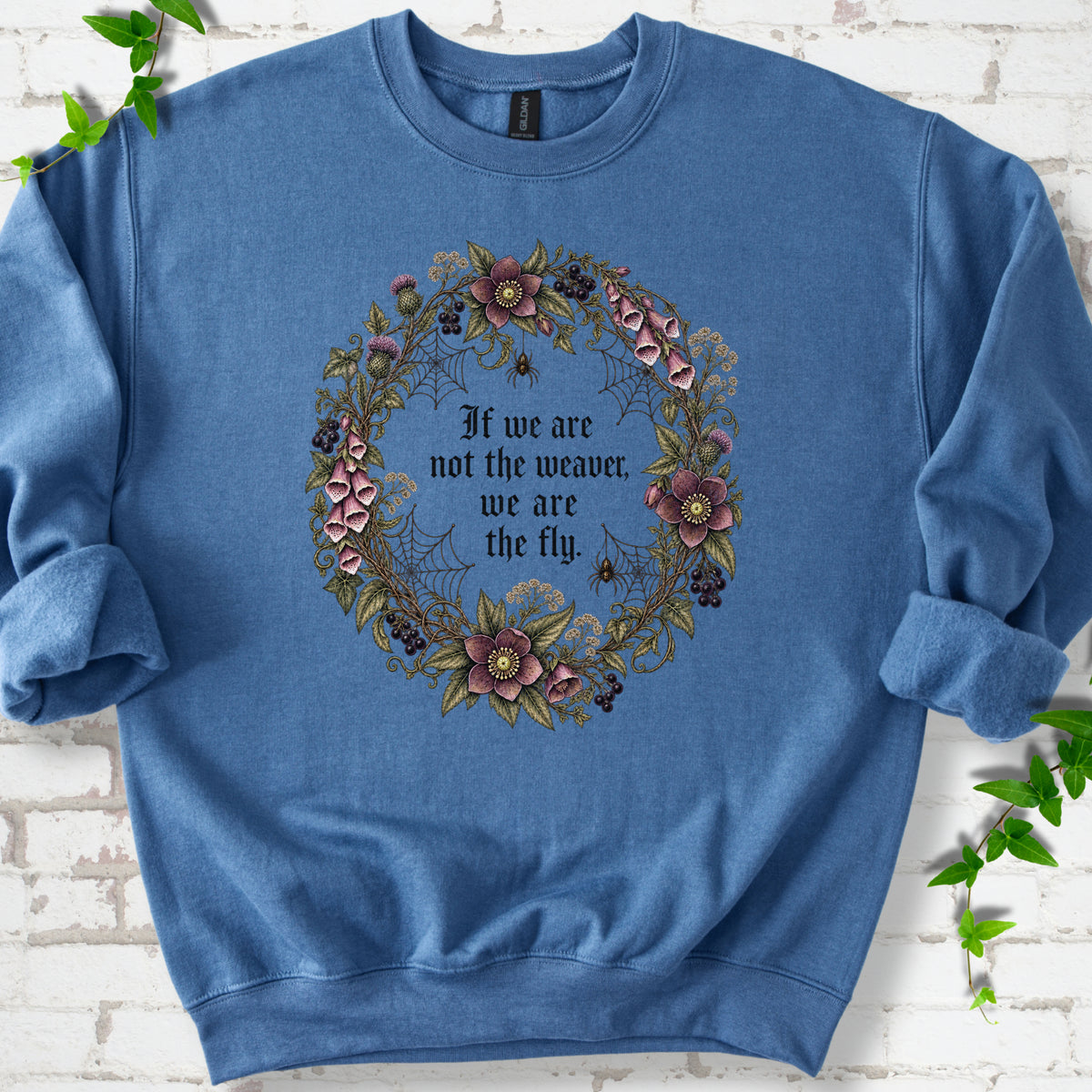 Indigo Blue sweatshirt flat-lay mockup with a gothic floral wreath, spider webs and "If we are not the weaver, we are the fly" lettering.