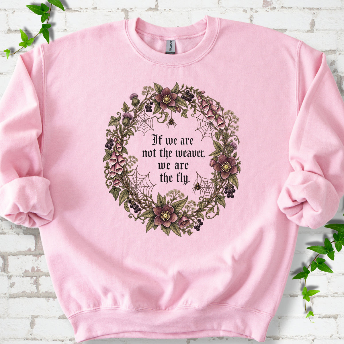 Light Pink sweatshirt flat-lay mockup with a gothic floral wreath, spider webs and "If we are not the weaver, we are the fly" lettering.