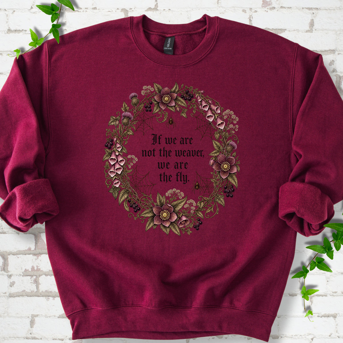Maroon sweatshirt flat-lay mockup with a gothic floral wreath, spider webs and "If we are not the weaver, we are the fly" lettering.