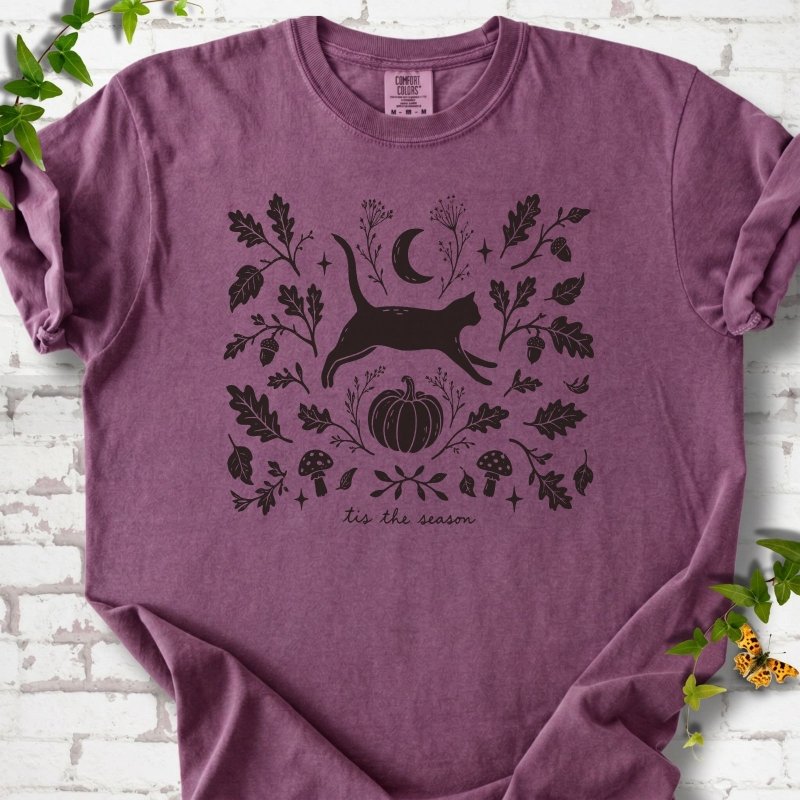 Berry Comfort Colors® T-shirt flat lay featuring a black cat leaping through autumn oak leaves, acorns, a moon, pumpkin and woodland mushrooms.