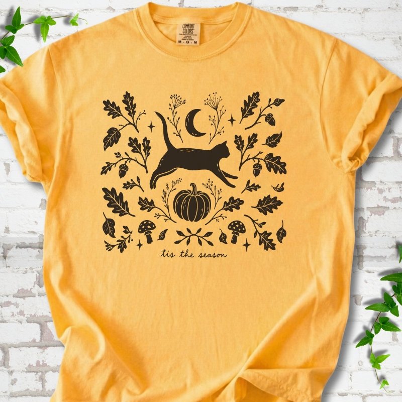 Citrus Comfort Colors® T-shirt flat lay featuring a black cat leaping through autumn oak leaves, acorns, a moon, pumpkin and woodland mushrooms.