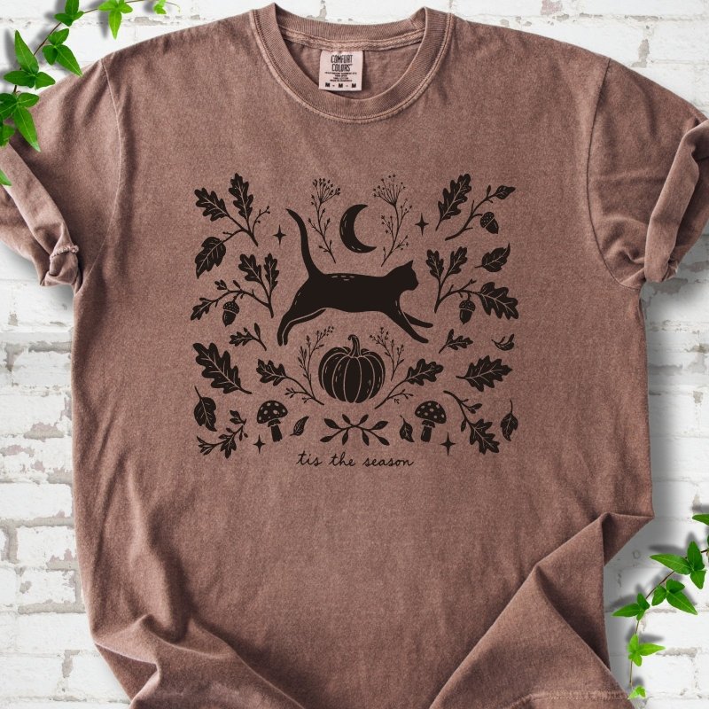 Espresso Comfort Colors® T-shirt flat lay featuring a black cat leaping through autumn oak leaves, acorns, a moon, pumpkin and woodland mushrooms.