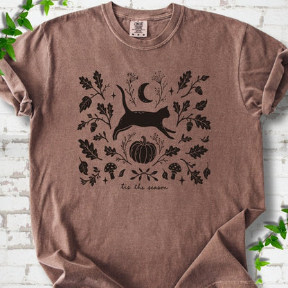 Espresso Comfort Colors® T-shirt flat lay featuring a black cat leaping through autumn oak leaves, acorns, a moon, pumpkin and woodland mushrooms.