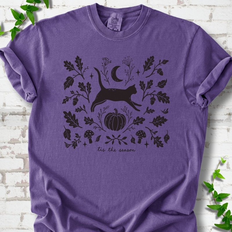 Grape Comfort Colors® T-shirt flat lay featuring a black cat leaping through autumn oak leaves, acorns, a moon, pumpkin and woodland mushrooms.