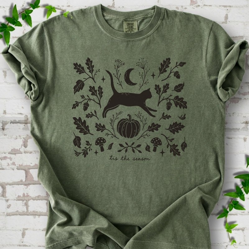 Moss Comfort Colors® T-shirt flat lay featuring a black cat leaping through autumn oak leaves, acorns, a moon, pumpkin and woodland mushrooms.