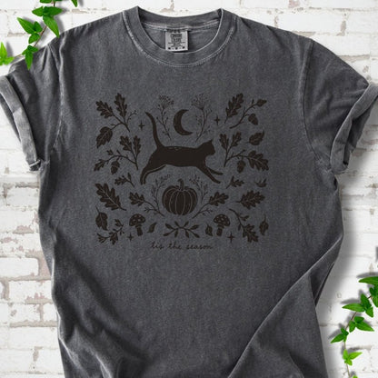 Pepper Comfort Colors® T-shirt flat lay featuring a black cat leaping through autumn oak leaves, acorns, a moon, pumpkin and woodland mushrooms.