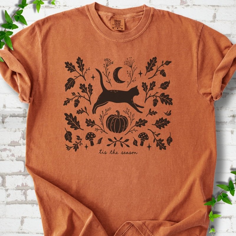 Yam Comfort Colors® T-shirt flat lay featuring a black cat leaping through autumn oak leaves, acorns, a moon, pumpkin and woodland mushrooms.