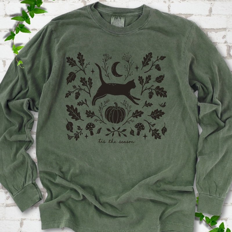 Hemp Comfort Colors® long sleeve T-shirt flat lay featuring a black cat leaping through autumn oak leaves, acorns, a moon, pumpkin and woodland mushrooms.