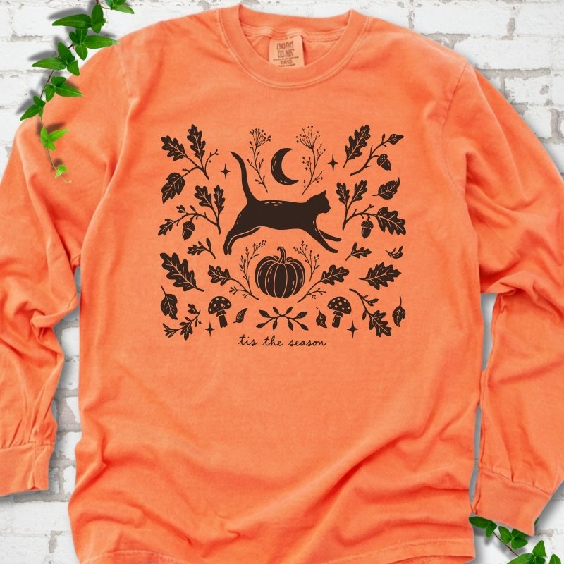 Melon Comfort Colors® long sleeve T-shirt flat lay featuring a black cat leaping through autumn oak leaves, acorns, a moon, pumpkin and woodland mushrooms.
