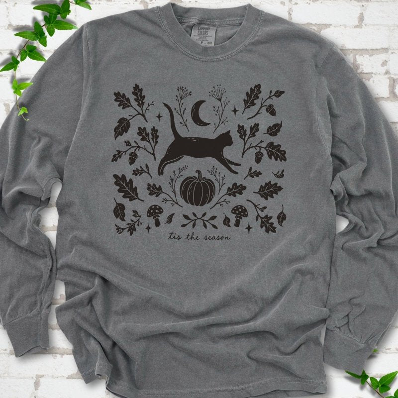 Pepper Comfort Colors® long sleeve T-shirt flat lay featuring a black cat leaping through autumn oak leaves, acorns, a moon, pumpkin and woodland mushrooms.