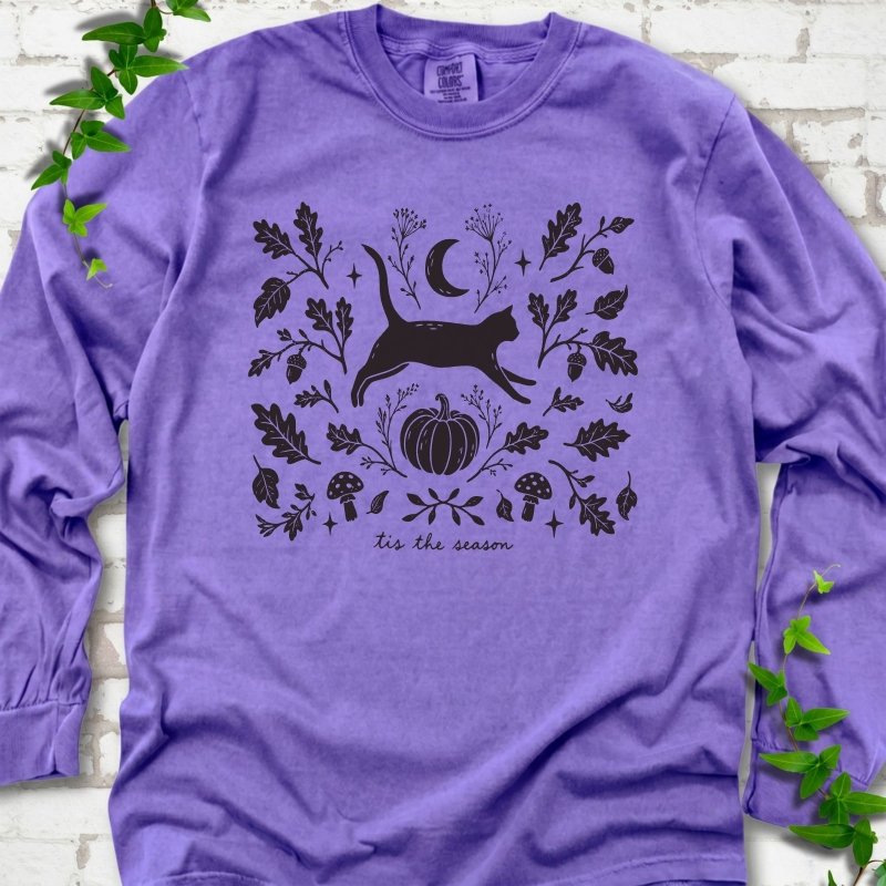 Violet Comfort Colors® long sleeve T-shirt flat lay featuring a black cat leaping through autumn oak leaves, acorns, a moon, pumpkin and woodland mushrooms.