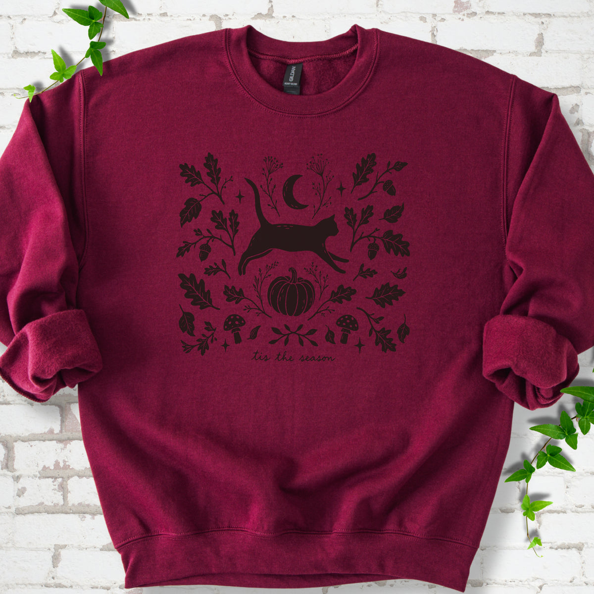 Maroon Gildan® 18000 crewneck sweatshirt flat lay featuring a black cat leaping among oak leaves, acorns, a crescent moon, pumpkin and mushrooms.