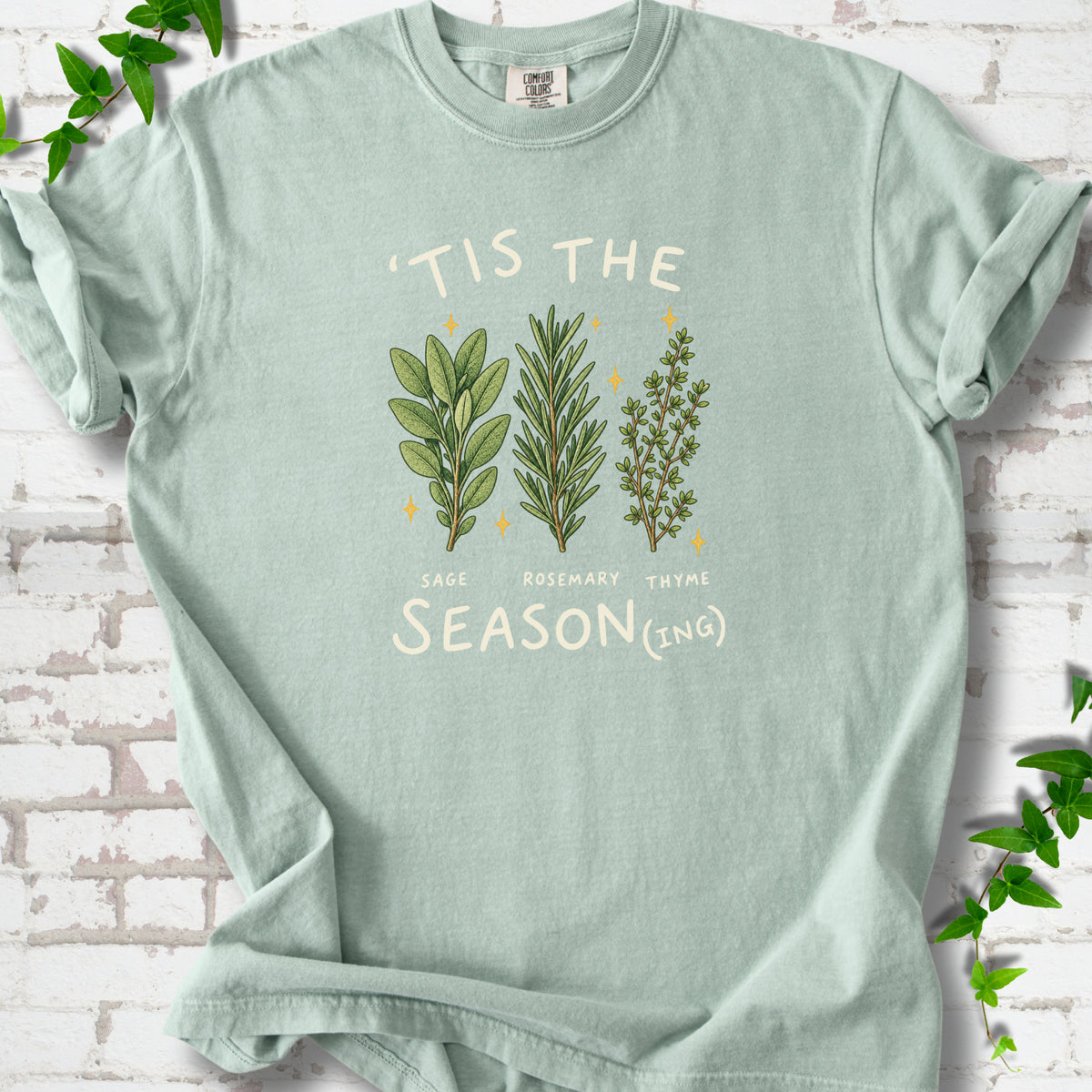 Bay T-shirt flat lay with ’Tis the Season(ing) lettering, sage, rosemary, thyme and small gold stars.