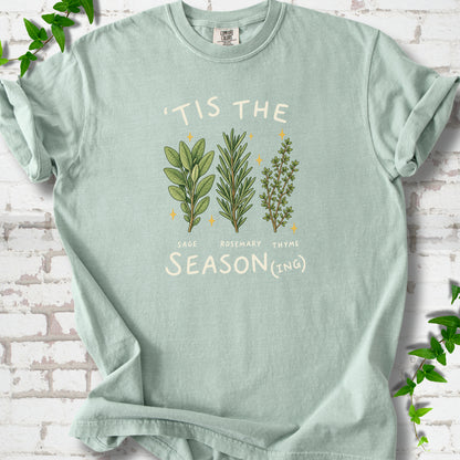 Bay T-shirt flat lay with ’Tis the Season(ing) lettering, sage, rosemary, thyme and small gold stars.