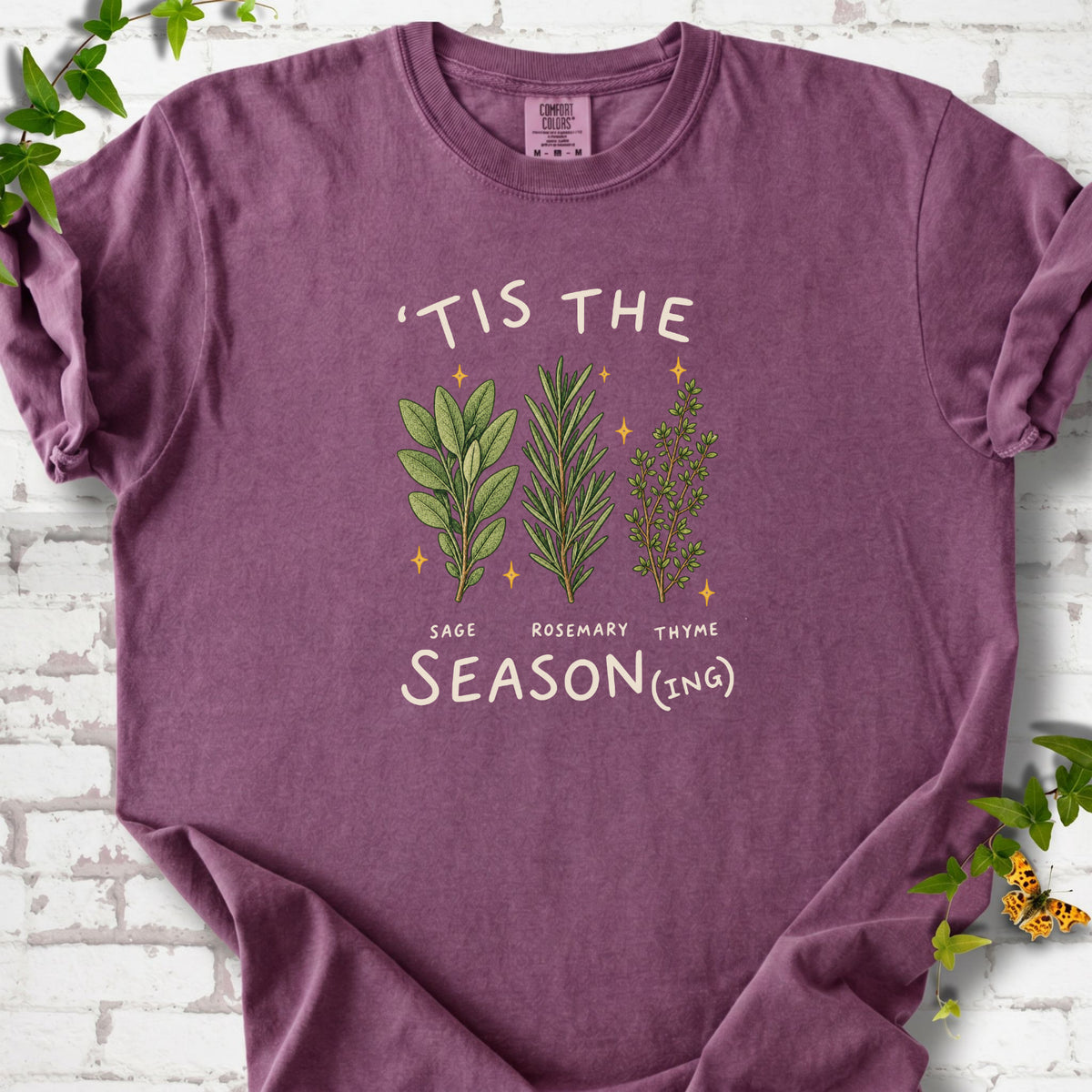 Berry T-shirt flat lay with ’Tis the Season(ing) lettering, sage, rosemary, thyme and small gold stars.