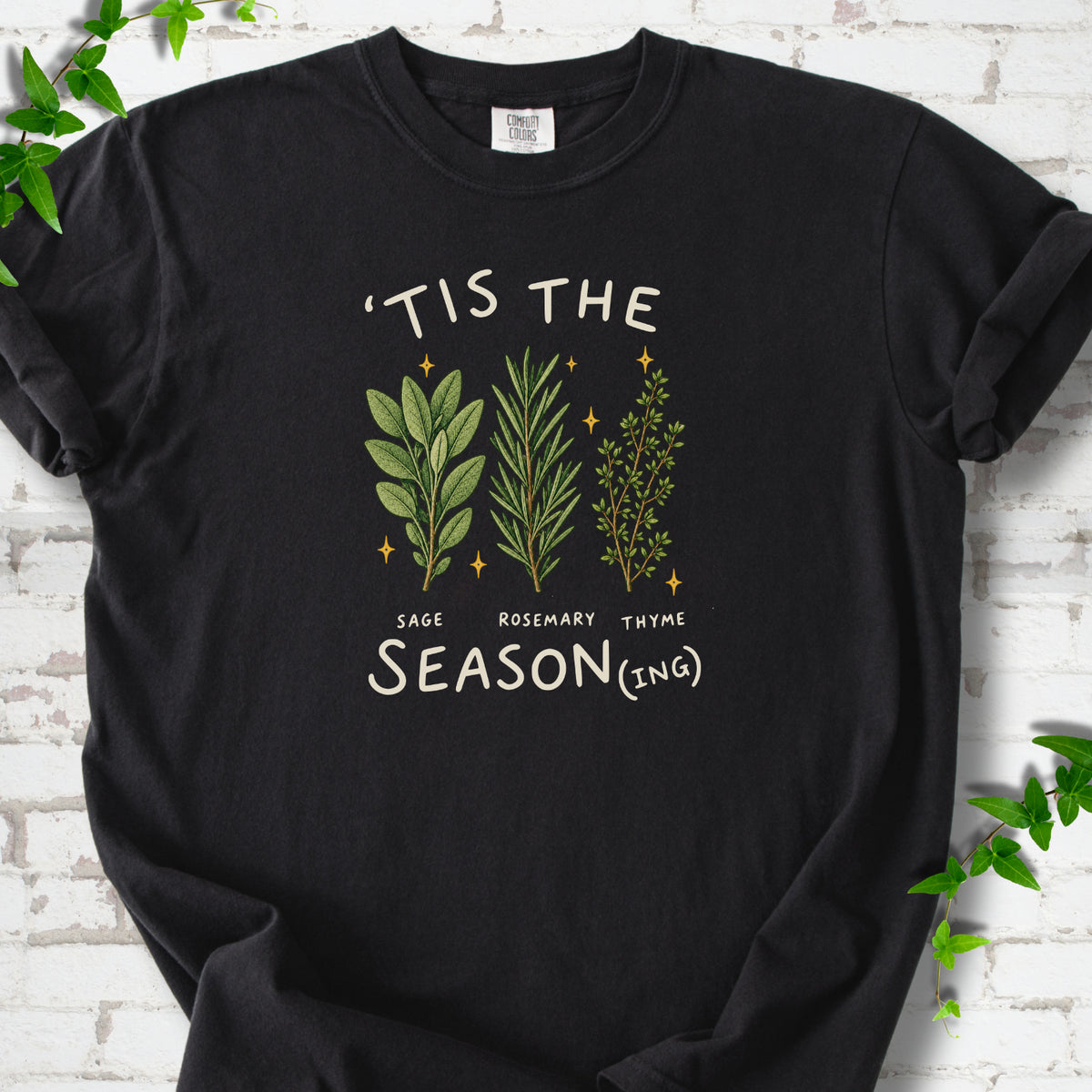 Black T-shirt flat lay with ’Tis the Season(ing) lettering, sage, rosemary, thyme and small gold stars.