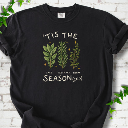 Black T-shirt flat lay with ’Tis the Season(ing) lettering, sage, rosemary, thyme and small gold stars.