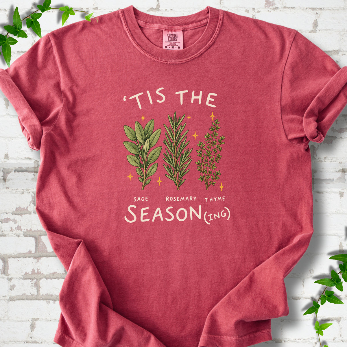 Crimson T-shirt flat lay with ’Tis the Season(ing) lettering, sage, rosemary, thyme and small gold stars.