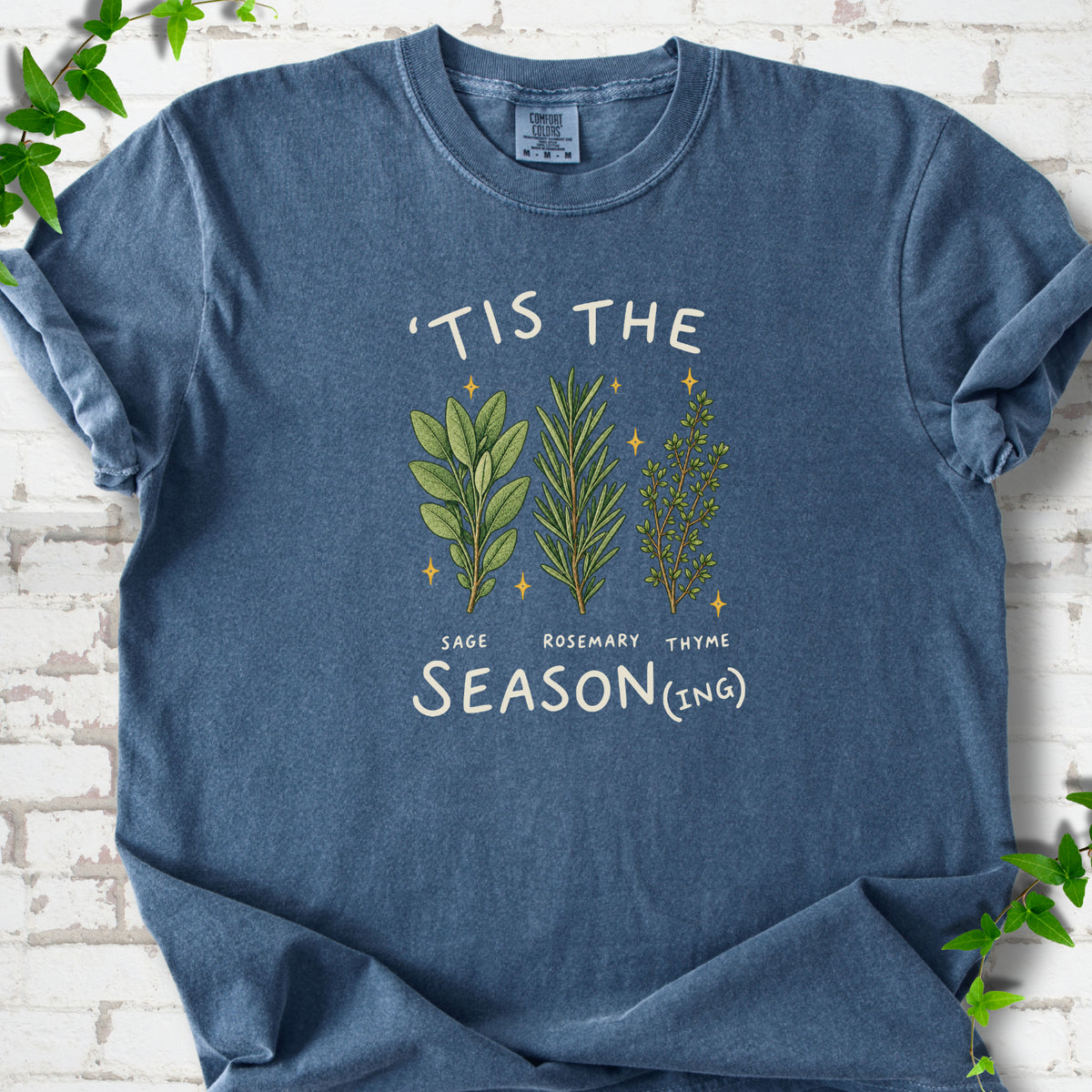 Denim T-shirt flat lay with ’Tis the Season(ing) lettering, sage, rosemary, thyme and small gold stars.