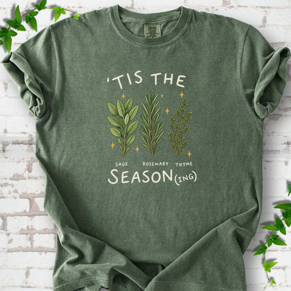 Hemp T-shirt flat lay with ’Tis the Season(ing) lettering, sage, rosemary, thyme and small gold stars.
