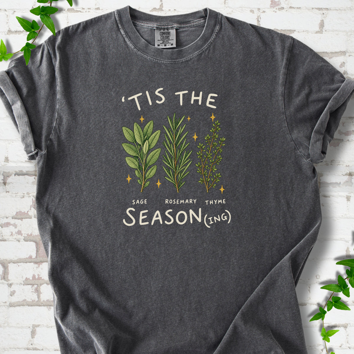 Pepper T-shirt flat lay with ’Tis the Season(ing) lettering, sage, rosemary, thyme and small gold stars.
