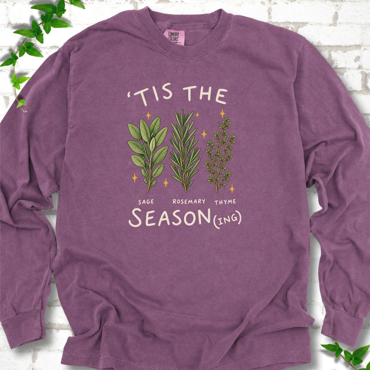 Berry long sleeve T-shirt flat lay with ’Tis the Season(ing) lettering, sage, rosemary, thyme and small gold stars.