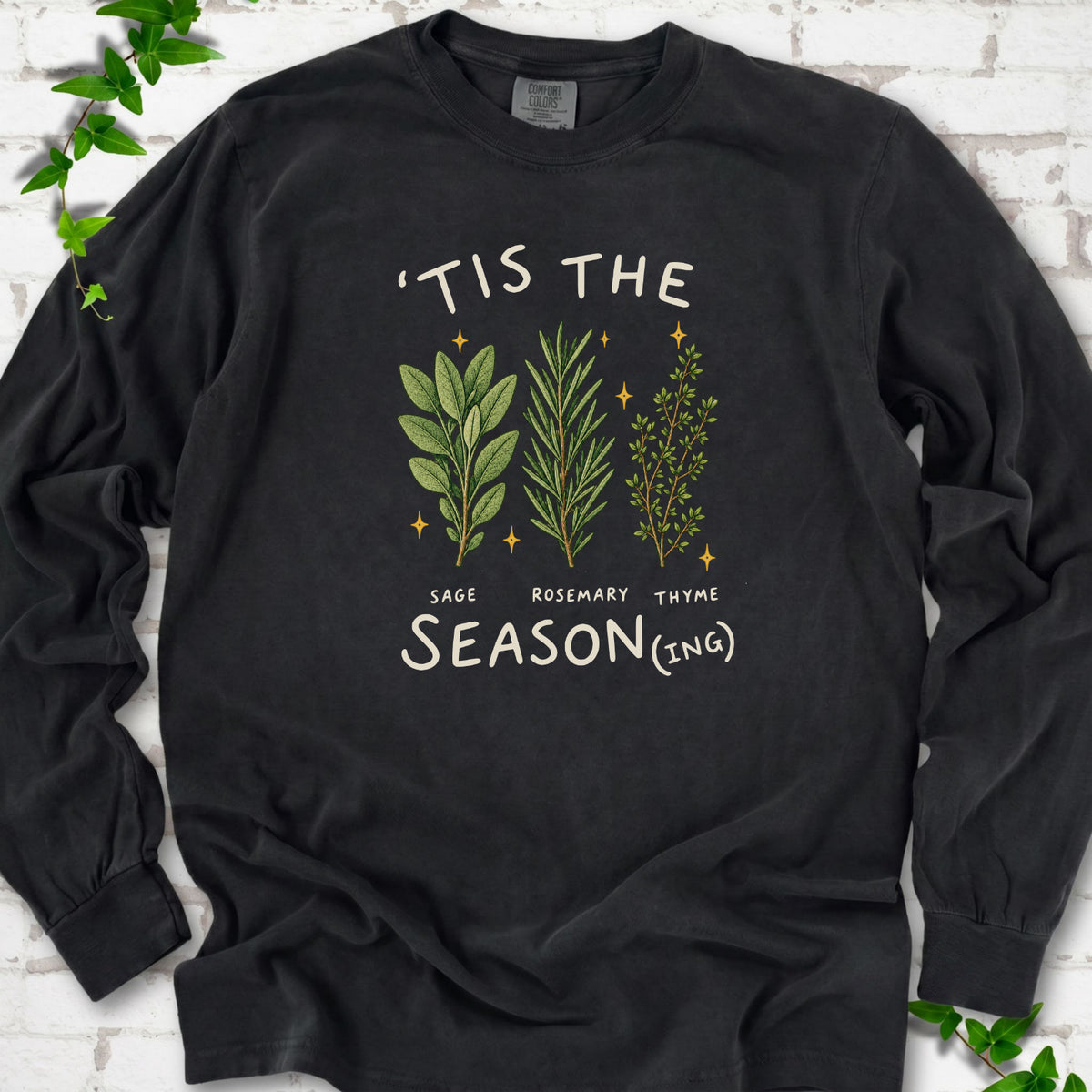 Black long sleeve T-shirt flat lay with ’Tis the Season(ing) lettering, sage, rosemary, thyme and small gold stars.