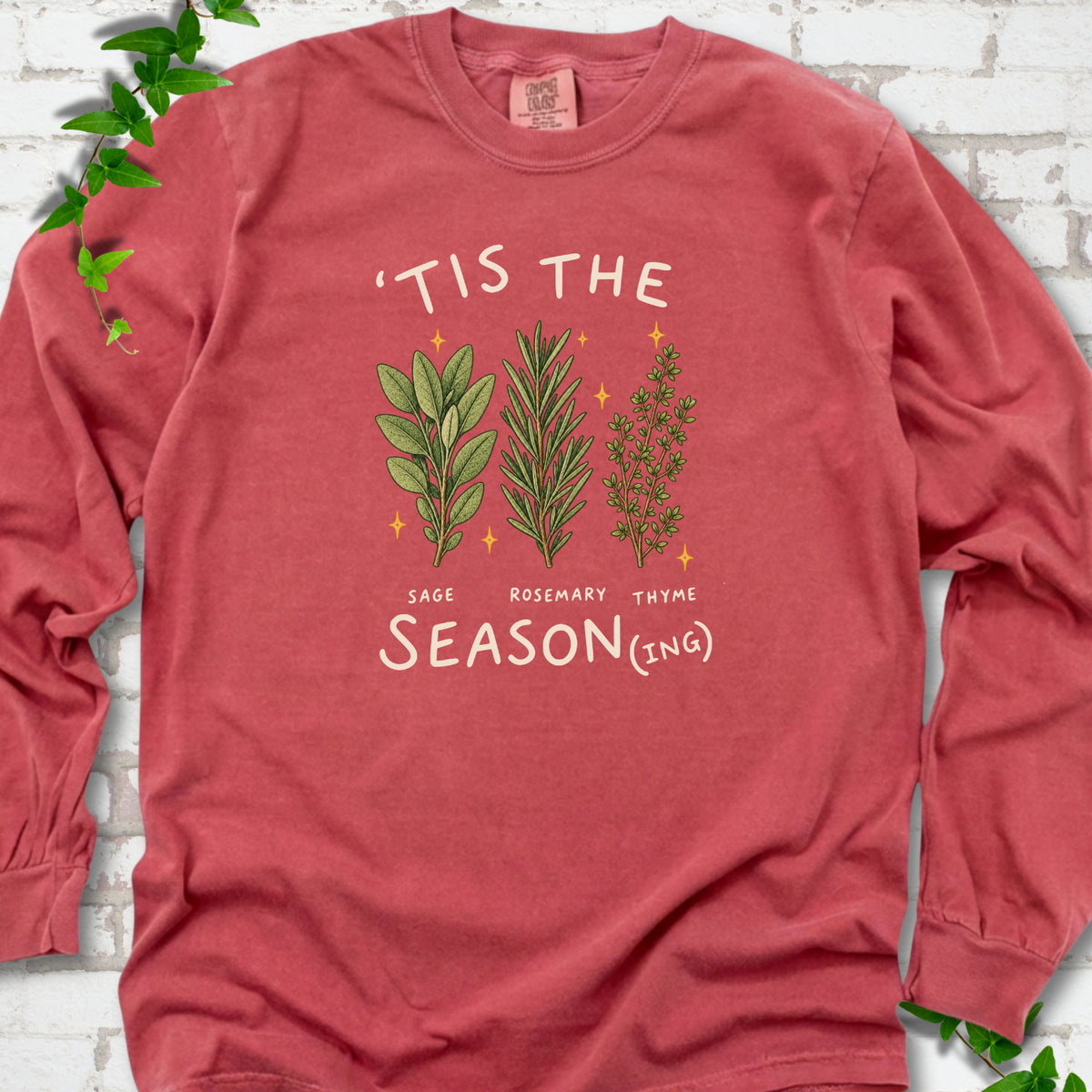 Crimson long sleeve T-shirt flat lay with ’Tis the Season(ing) lettering, sage, rosemary, thyme and small gold stars.