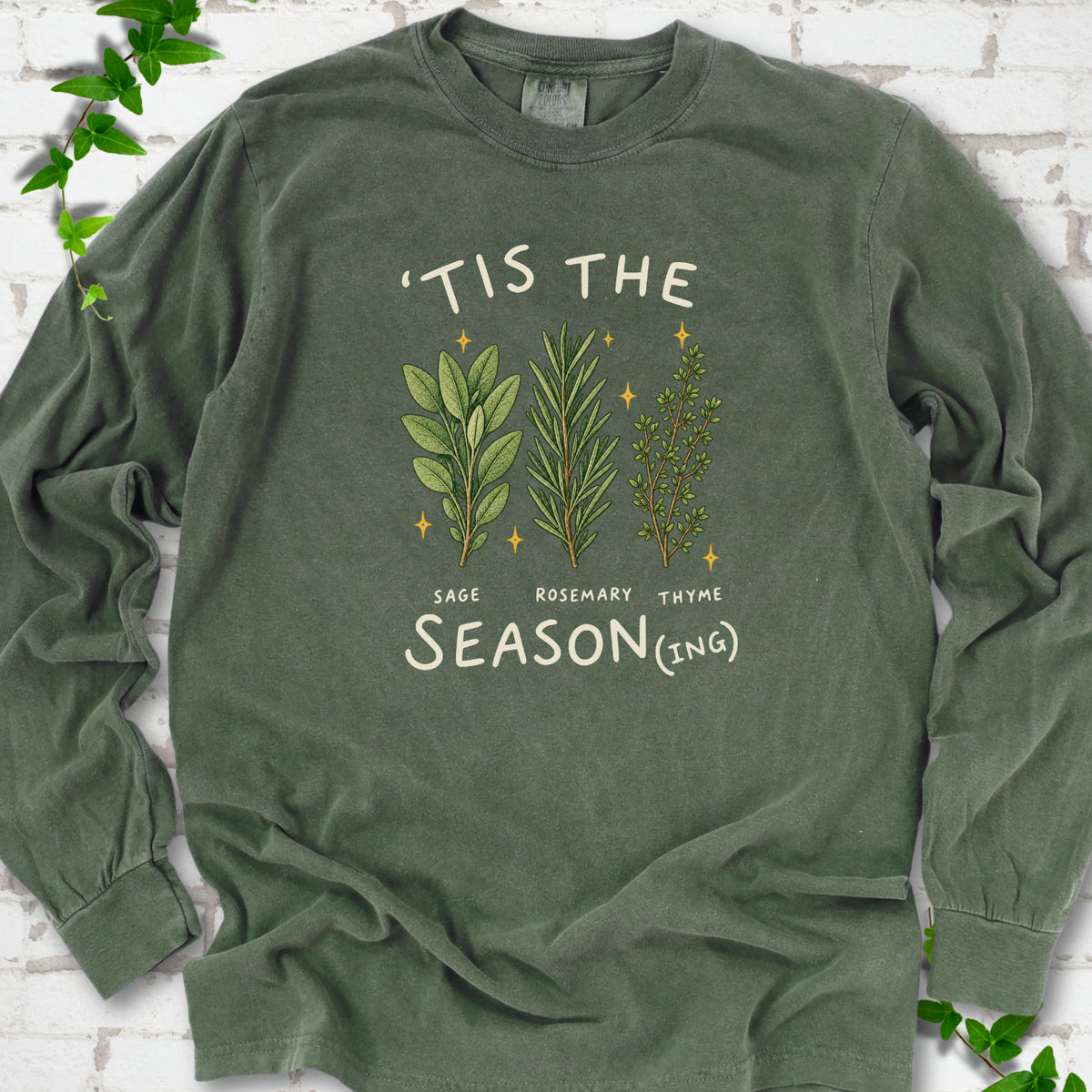 Hemp long sleeve T-shirt flat lay with ’Tis the Season(ing) lettering, sage, rosemary, thyme and small gold stars.