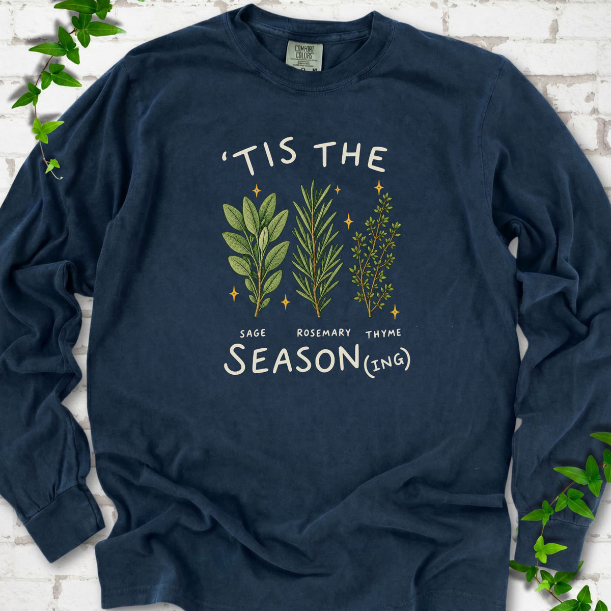 Midnight long sleeve T-shirt flat lay with ’Tis the Season(ing) lettering, sage, rosemary, thyme and small gold stars.