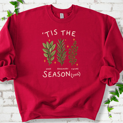 Cardinal Red crewneck sweatshirt flat lay with ’Tis the Season(ing) lettering, sage, rosemary, thyme and small gold stars.