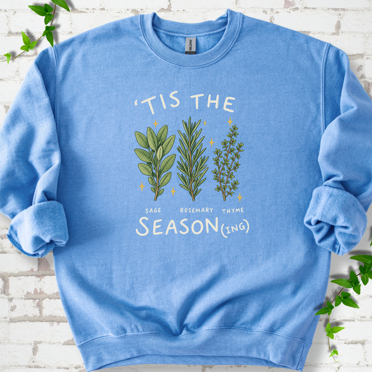 Carolina Blue crewneck sweatshirt flat lay with ’Tis the Season(ing) lettering, sage, rosemary, thyme and small gold stars.