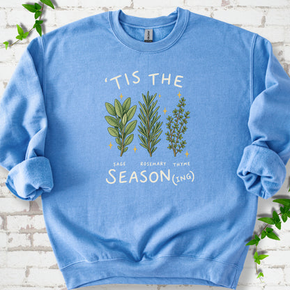 Carolina Blue crewneck sweatshirt flat lay with ’Tis the Season(ing) lettering, sage, rosemary, thyme and small gold stars.