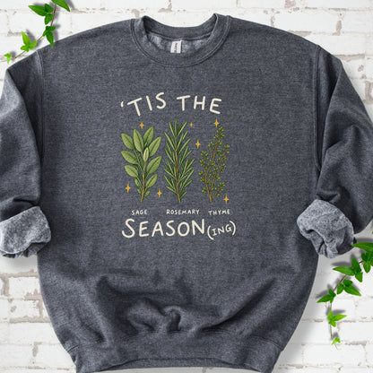 Dark Heather crewneck sweatshirt flat lay with ’Tis the Season(ing) lettering, sage, rosemary, thyme and small gold stars.