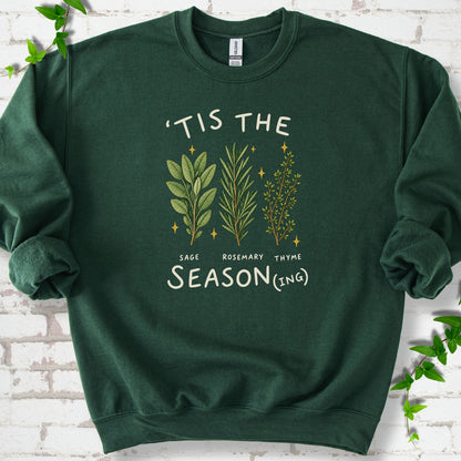 Forest Green crewneck sweatshirt flat lay with ’Tis the Season(ing) lettering, sage, rosemary, thyme and small gold stars.