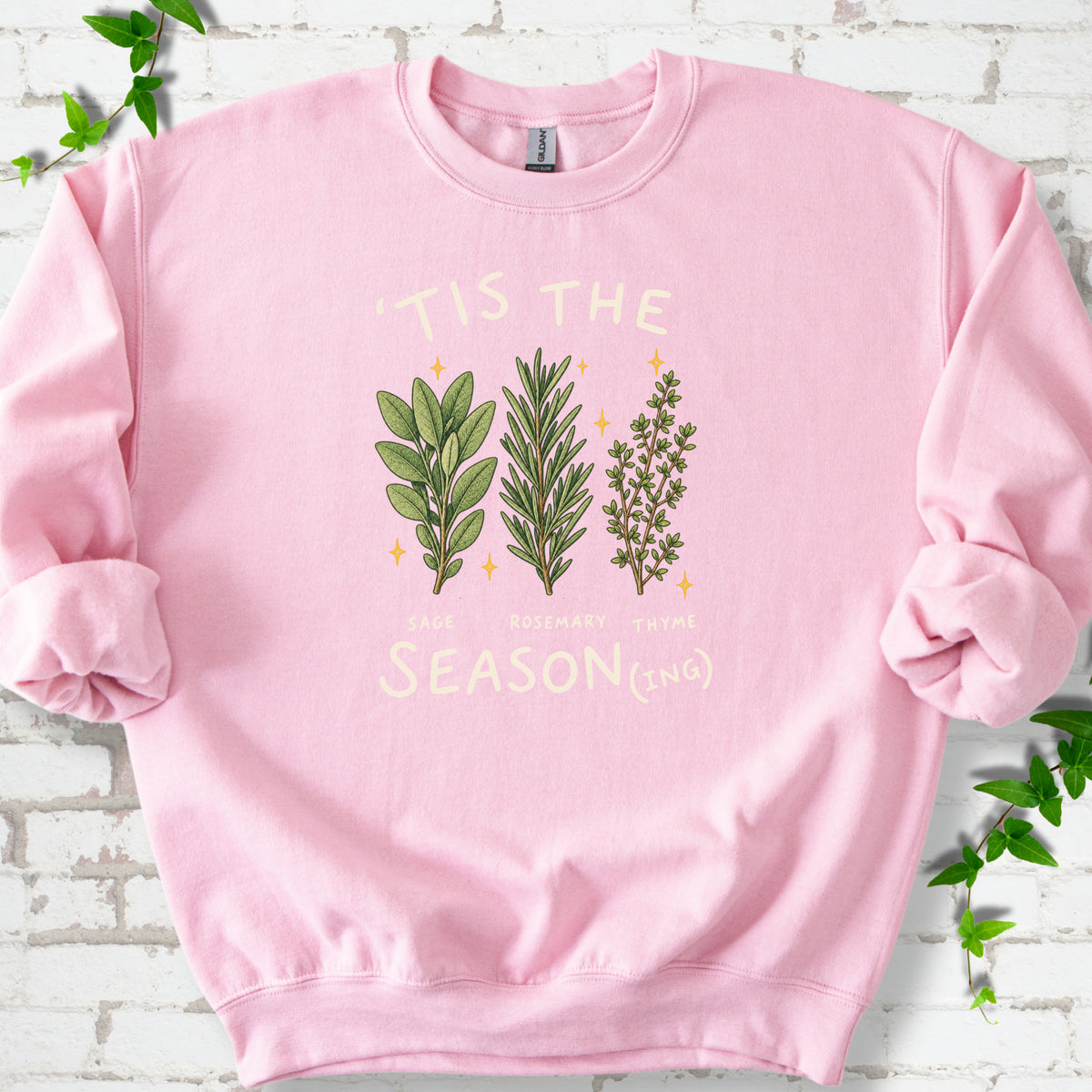 Light Pink crewneck sweatshirt flat lay with ’Tis the Season(ing) lettering, sage, rosemary, thyme and small gold stars.