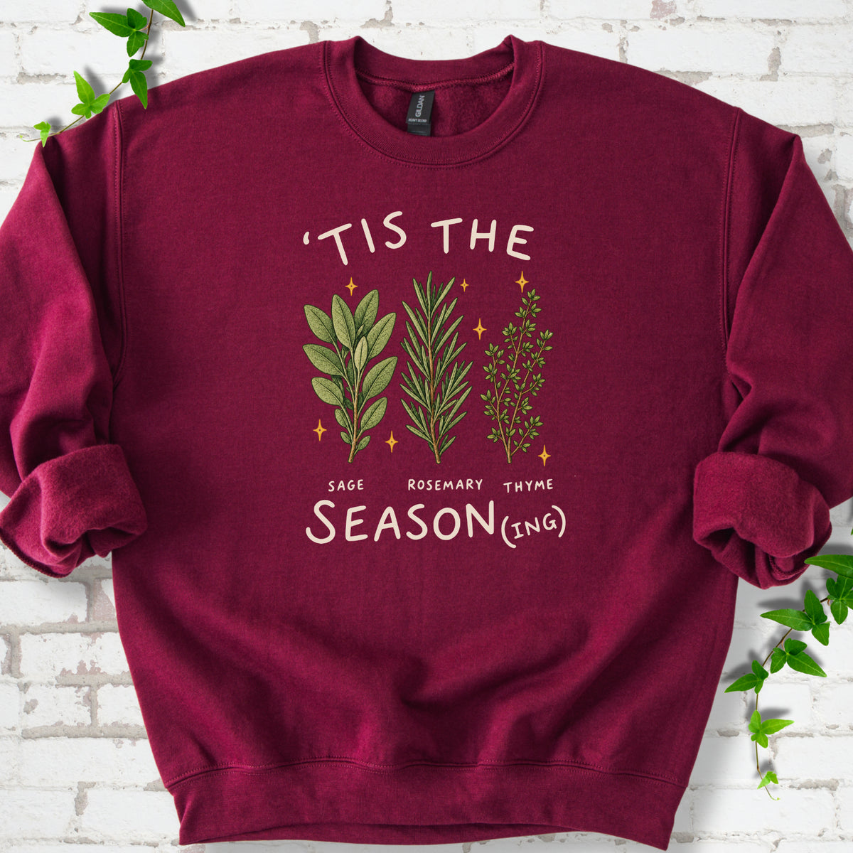 Maroon crewneck sweatshirt flat lay with ’Tis the Season(ing) lettering, sage, rosemary, thyme and small gold stars.