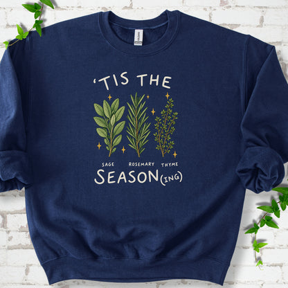 Navy crewneck sweatshirt flat lay with ’Tis the Season(ing) lettering, sage, rosemary, thyme and small gold stars.