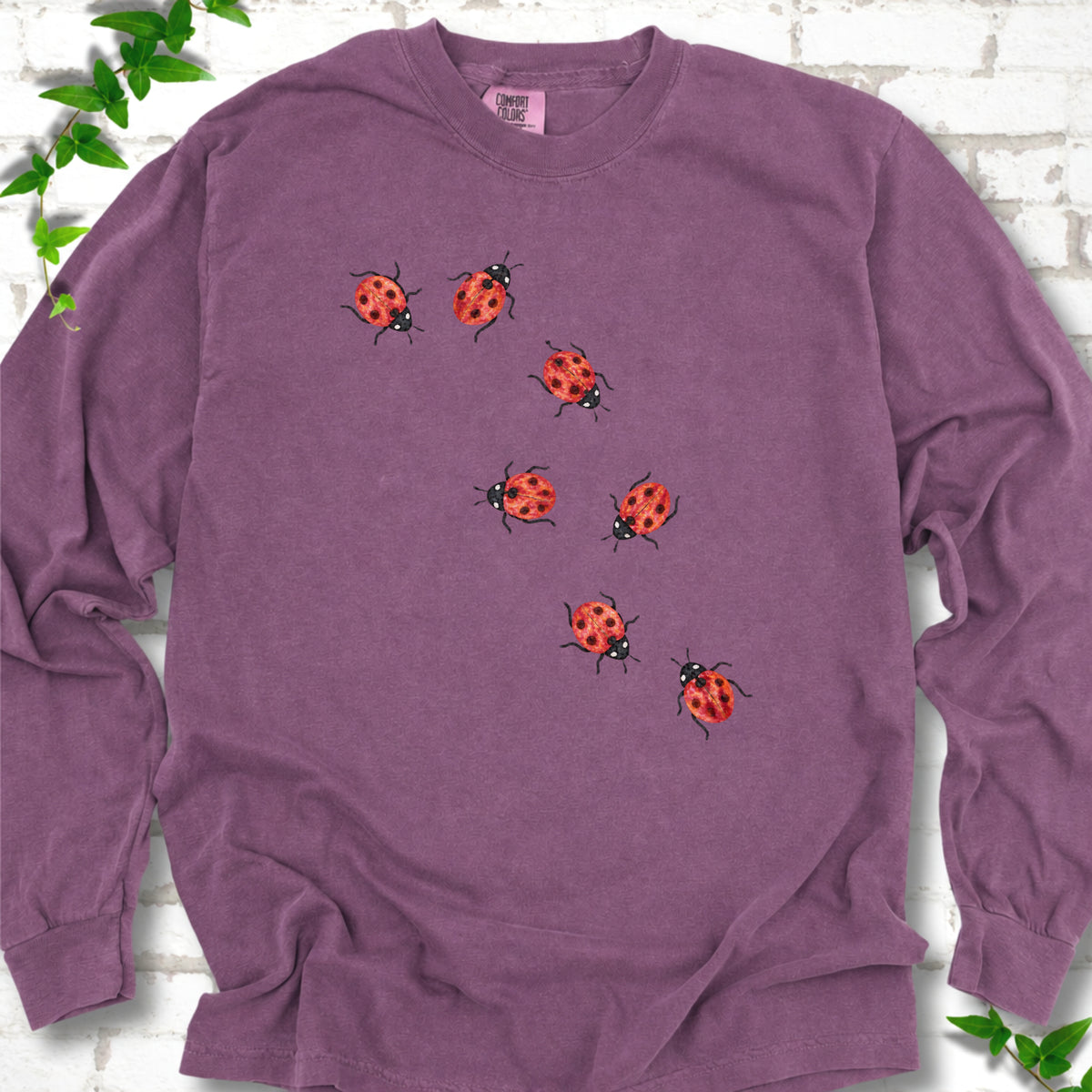 Berry purple long sleeve T-shirt with seven small red ladybugs walking in a diagonal trail across the chest, flat lay on white brick with ivy