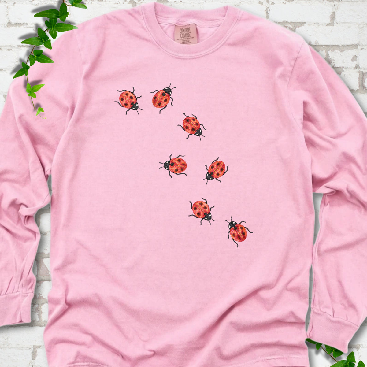 Blossom pink long sleeve T-shirt with seven small red ladybugs walking in a diagonal trail across the chest, flat lay on white brick with ivy
