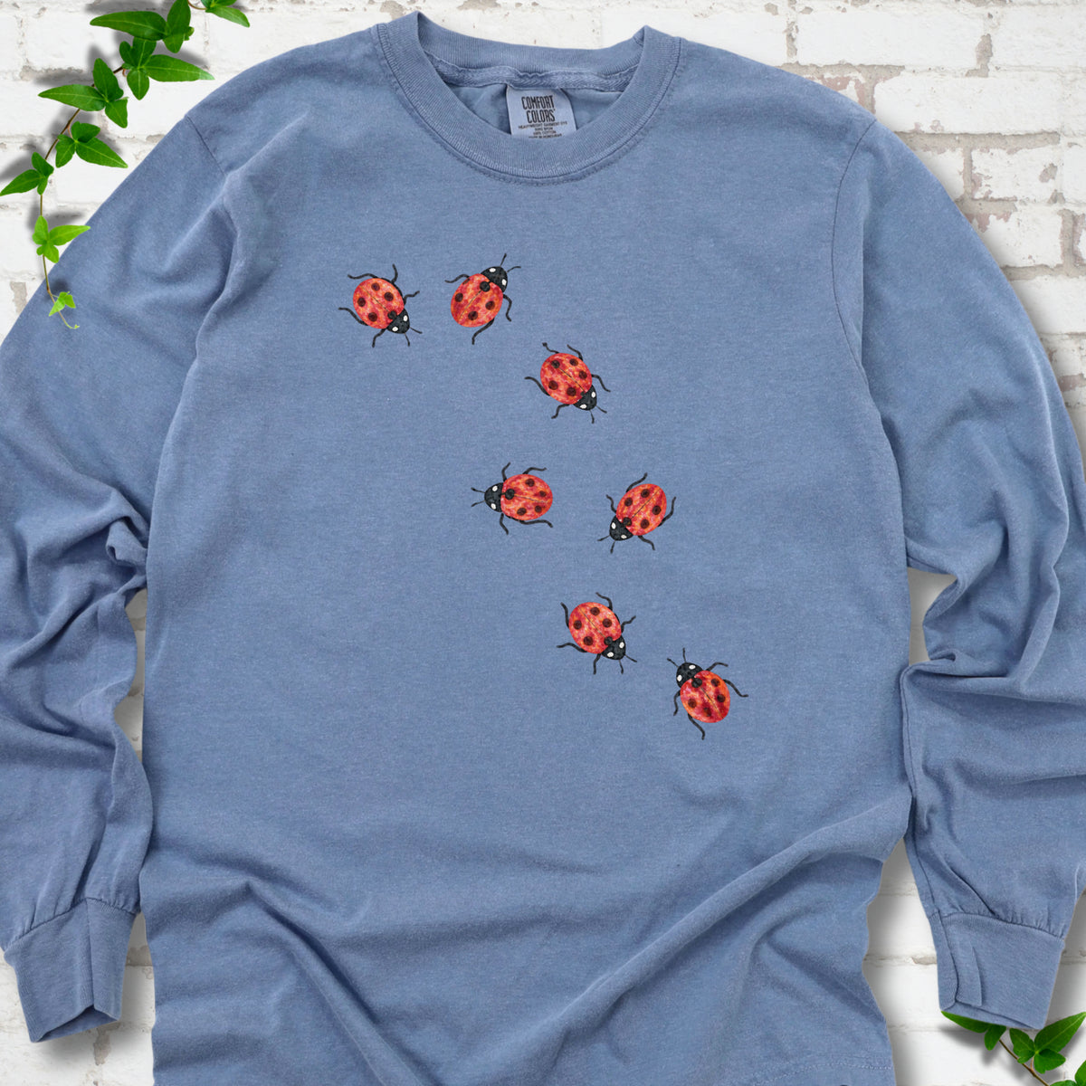 Blue Jean light blue long sleeve T-shirt with seven small red ladybugs walking in a diagonal trail across the chest, flat lay on white brick with ivy