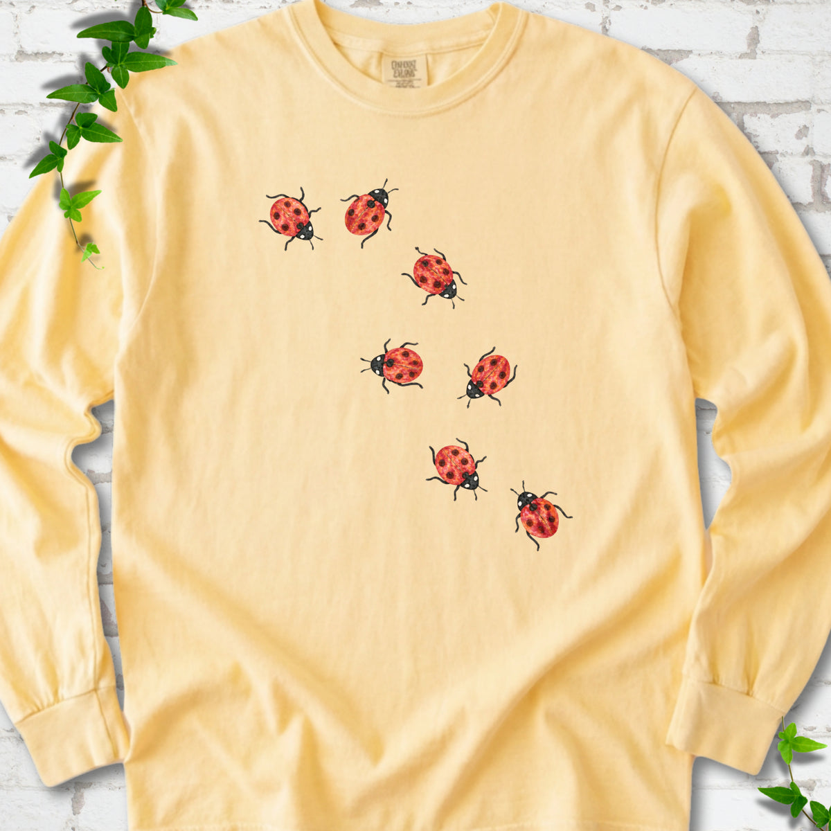 Butter yellow long sleeve T-shirt with seven small red ladybugs walking in a diagonal trail across the chest, flat lay on white brick with ivy