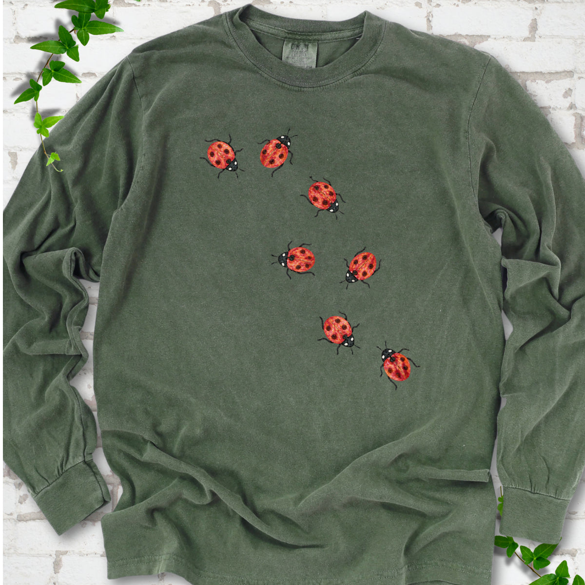 Hemp green long sleeve T-shirt with seven small red ladybugs walking in a diagonal trail across the chest, flat lay on white brick with ivy