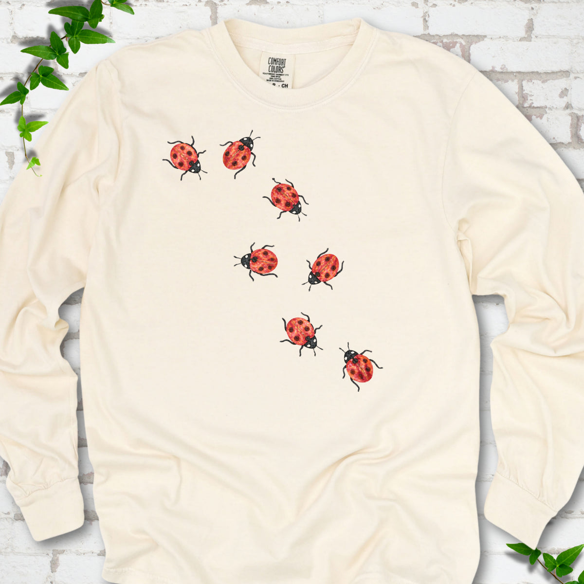 Ivory cream long sleeve T-shirt with seven small red ladybugs walking in a diagonal trail across the chest, flat lay on white brick with ivy