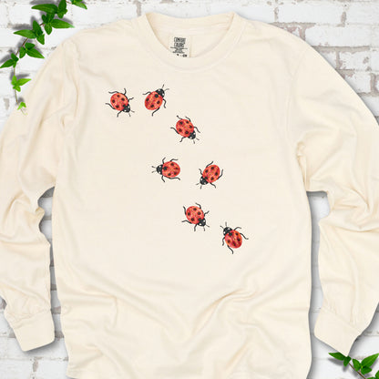 Ivory cream long sleeve T-shirt with seven small red ladybugs walking in a diagonal trail across the chest, flat lay on white brick with ivy