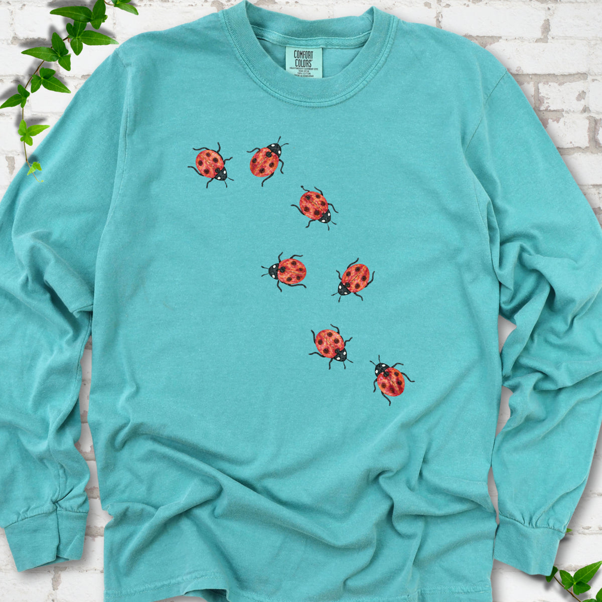 Seafoam teal long sleeve T-shirt with seven small red ladybugs walking in a diagonal trail across the chest, flat lay on white brick with ivy