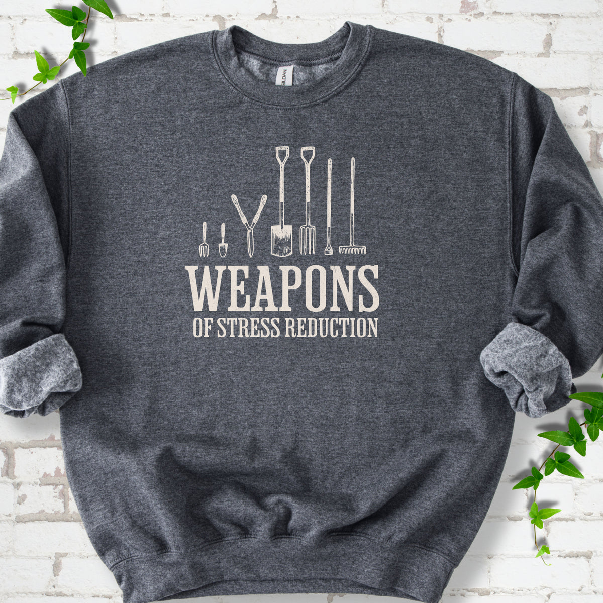Dark Heather Gildan® 18000 crewneck sweatshirt flat lay featuring garden tools and the words “Weapons of Stress Reduction.”